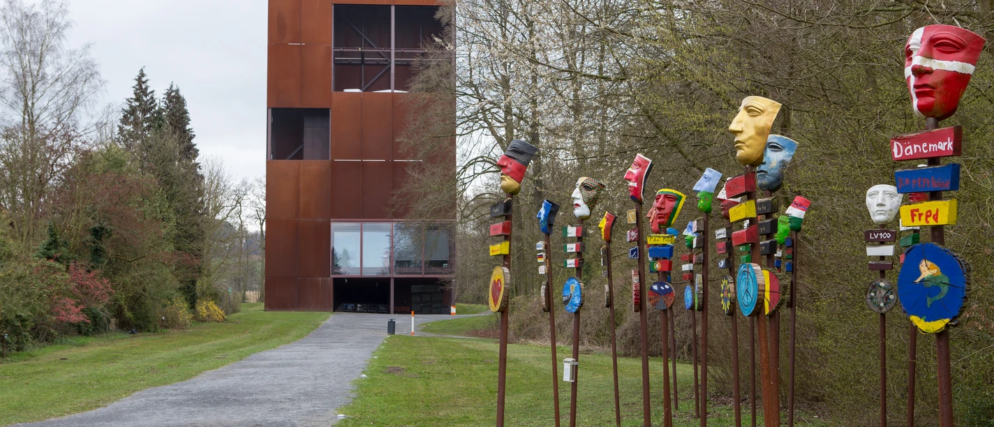 Varusschlacht im Osnabrücker Land - Museum und Park Kalkriese Rusty tower next to colorful, painted mask poles on a path through trees.