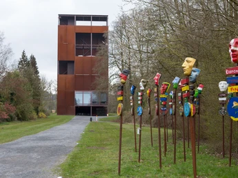 Varusschlacht im Osnabrücker Land - Museum und Park Kalkriese Rostiger Turm neben bunten, bemalten Maskenpfählen auf einem Weg durch Bäume.Rusty tower next to colorful, painted mask poles on a path through trees.Rustent tårn ved siden af farverige, malede maskestolper på en sti gennem træerne.Roestige toren naast kleurrijke, beschilderde maskerpalen op een pad tussen bomen.