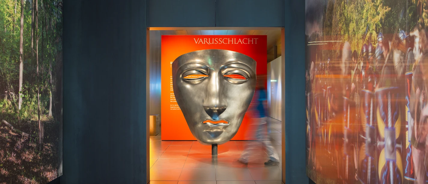 Varusschlacht im Osnabrücker Land - Museum und Park Kalkriese Large silver mask in the Varus Battle Museum with visitors in the hallway.