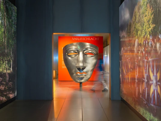 Varusschlacht im Osnabrücker Land - Museum und Park Kalkriese Große silberne Maske im Varusschlacht-Museum mit Besucher im Flur.Large silver mask in the Varus Battle Museum with visitors in the hallway.Stor sølvmaske i Varus Battle Museum med besøgende i korridoren.Groot zilveren masker in het Varusslagmuseum met bezoekers in de gang.
