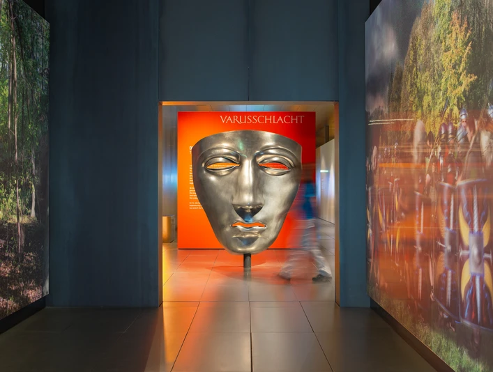 Varusschlacht im Osnabrücker Land - Museum und Park Kalkriese Große silberne Maske im Varusschlacht-Museum mit Besucher im Flur.Large silver mask in the Varus Battle Museum with visitors in the hallway.Stor sølvmaske i Varus Battle Museum med besøgende i korridoren.Groot zilveren masker in het Varusslagmuseum met bezoekers in de gang.