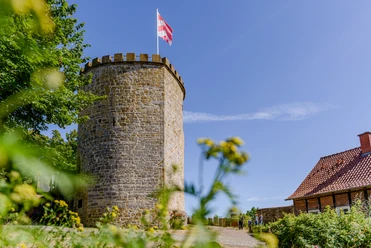 Runder Burgturm aus Stein mit Zinnen und wehender rot-weißer Fahne bei klarem Himmel.