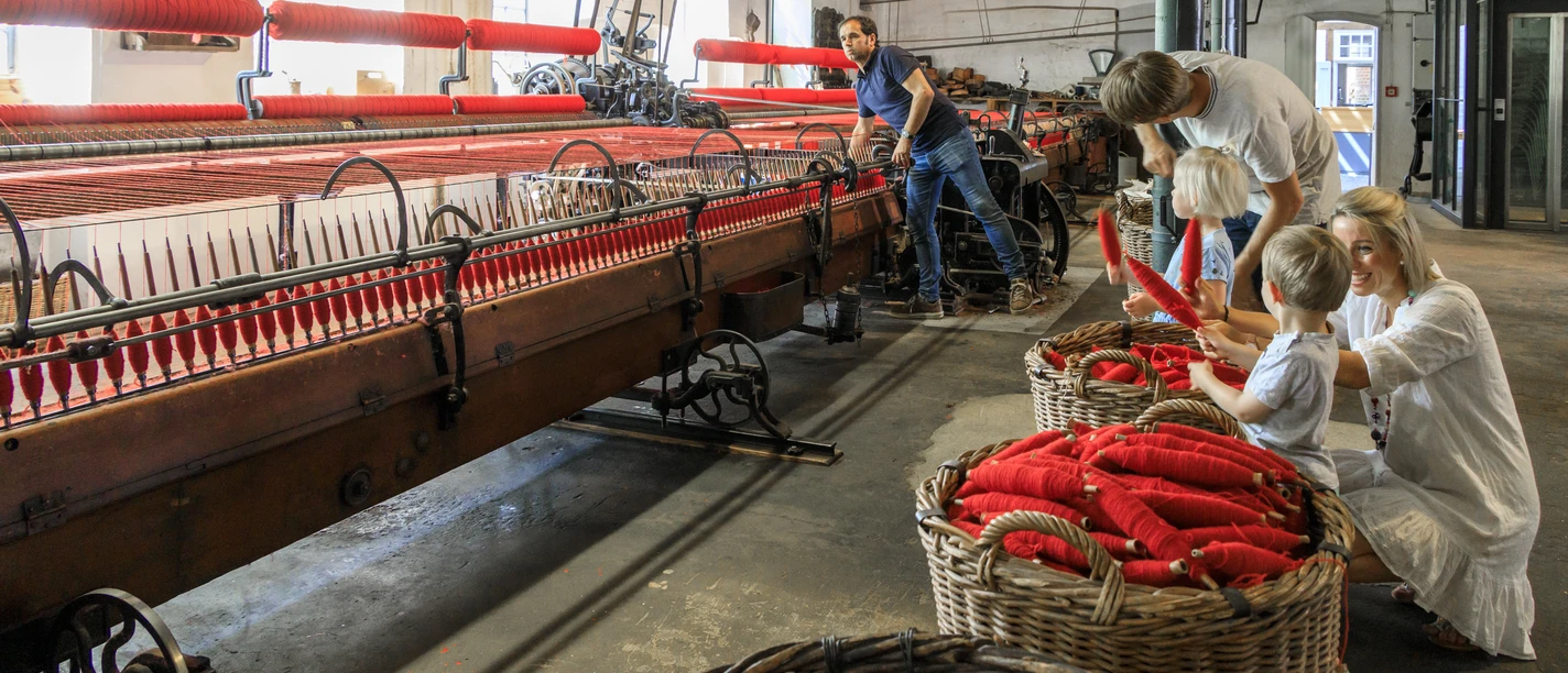 bramsche-tuchmacher-museum(c)tol-foto-jurjen-drenth (11).JPG Historische Weberei mit Besuchern, roten Garnspulen, alten Maschinen in einem lichtdurchfluteten Raum.