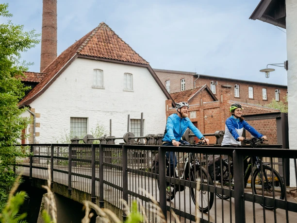 Am Tuchmacher Museum in Bramsche Twee fietsers steken een brug over voor een historisch gebouw met een schoorsteen.