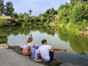 Alfsee Ferien- und Erlebnispark Familie am Teich auf Steinmauer sitzend, umgeben von Bäumen, beobachtet das Wasser.