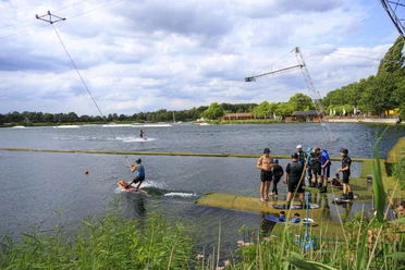 alfsee-ferienpark-germanenland(c)tol-foto-jurjen-drenth (47).JPG Wasserskifahrer an einer Seilbahn auf einem See, umgeben von grüner Natur.