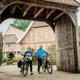 artland-hofcafe©tol-foto-christoph-steinweg (10).jpg Zwei Radfahrer stehen unter einem hölzernen Torbogen, umgeben von Fachwerkhäusern.