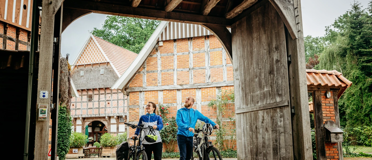artland-hofcafe©tol-foto-christoph-steinweg (10).jpg Zwei Radfahrer stehen unter einem hölzernen Torbogen, umgeben von Fachwerkhäusern.