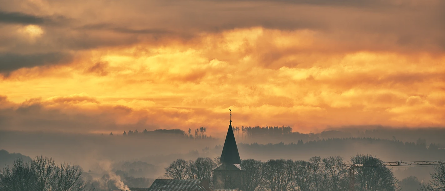 hagen-atw-cc0-domenic-schlinge.jpeg Dorf mit Kirchturm vor spektakulärem Sonnenuntergang und Nebel in der ländlichen Silhouette.