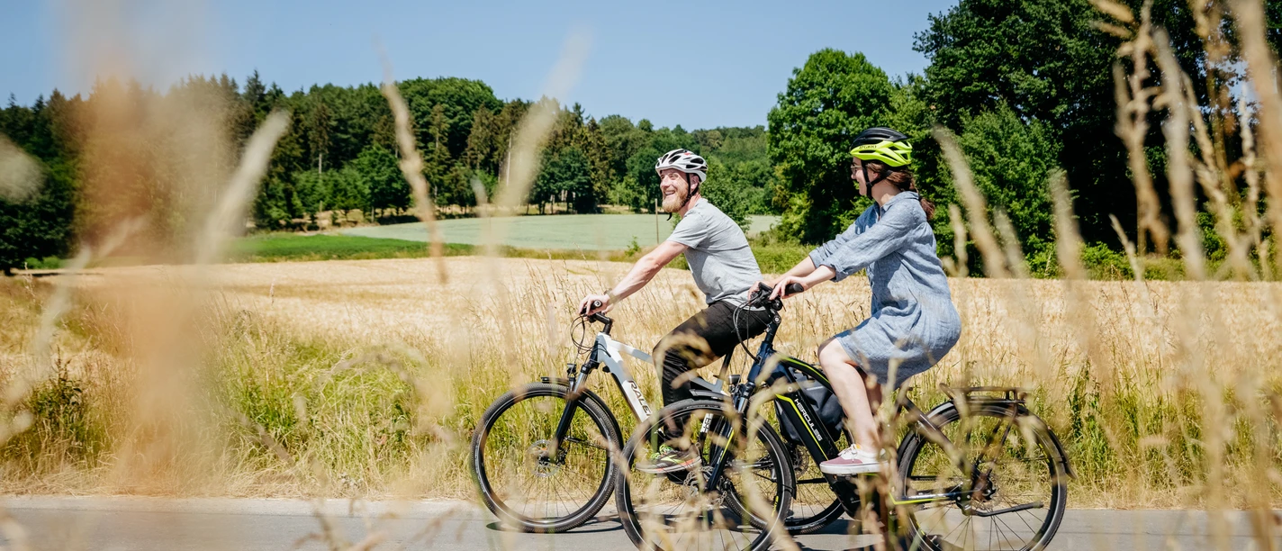 Radfahren im Osnabrücker Land Zwei Radfahrer mit Helmen fahren durch eine sonnige Landschaft mit Wiesen und Wald.