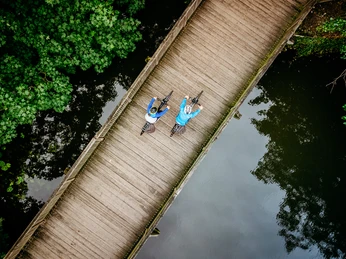 Mit dem Rad durch das Osnabrücker Land Zwei Radfahrer auf einer Holzbrücke über einem ruhigen Fluss, umgeben von grünen Bäumen.Two cyclists on a wooden bridge over a quiet river, surrounded by green trees.To cyklister på en træbro over en stille flod, omgivet af grønne træer.Twee fietsers op een houten brug over een rustige rivier, omringd door groene bomen.