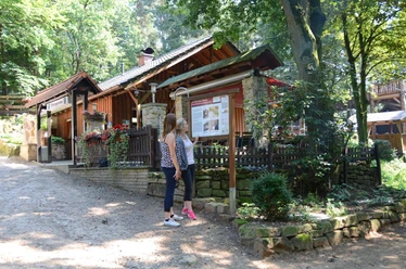 Ibbenbüren-Almhütte-1600.jpg Zwei Frauen stehen vor einer Informationstafel vor einer Holzhütte im Wald an einem sonnigen Tag.
