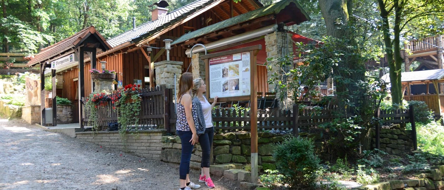 Ibbenbüren-Almhütte-1600.jpg Zwei Frauen stehen vor einer Informationstafel vor einer Holzhütte im Wald an einem sonnigen Tag.