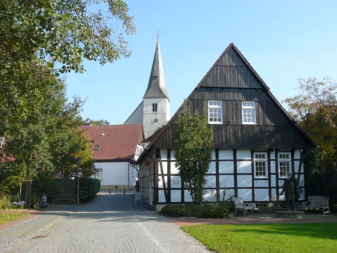 Fachwerkhaus mit weißer Fassade und dunklen Holzbalken, im Hintergrund eine Kirche mit hoher Turmspitze.