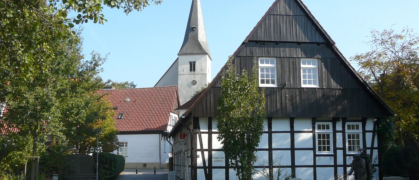 Lienen_Haus des Gastes_Foto_Gemeinde Lienen.JPG Fachwerkhaus mit weißer Fassade und dunklen Holzbalken, im Hintergrund eine Kirche mit hoher Turmspitze.