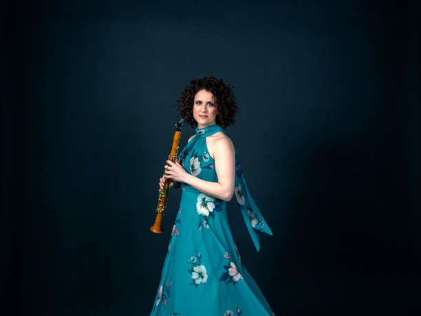 A woman in a teal floral dress holding a clarinet against a dark backdrop.
