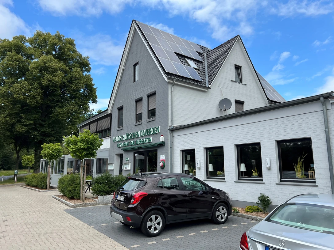 Ein zweistöckiges Gebäude mit weißen Wänden, grauem Dach und Solarpanelen, im Vordergrund geparkte Autos.