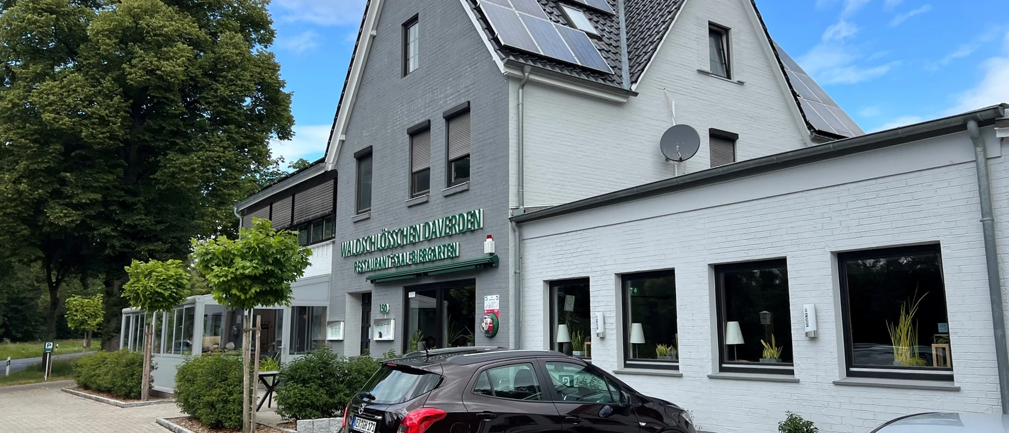 Een gebouw van twee verdiepingen met witte muren, een grijs dak en zonnepanelen, met geparkeerde auto's op de voorgrond.