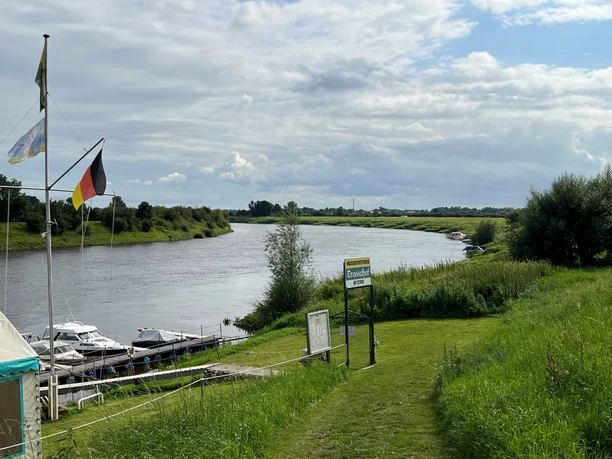Am Ufer eines breiten Flusses liegt ein kleiner Hafen mit Segelbooten, teils unter bewölktem Himmel.
