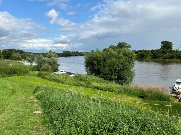 Weser bei Drosselhof Grüner Uferstreifen mit Bäumen und Booten an der ruhig fließenden Weser unter bewölktem Himmel.