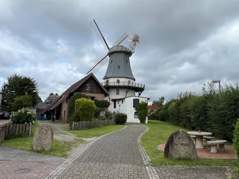 Historische Windmühle in Etelsen mit grüner Umgebung und benachbartem Backsteingebäude unter wolkigem Himmel.