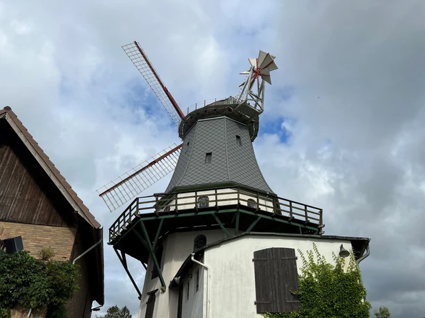 Historische Windmühle Etelsen mit grauer Fassade und rotem Flügelkreuz unter bewölktem Himmel.