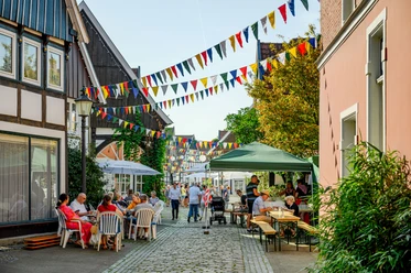 Bunt beflaggte Kopfsteinpflasterstraße mit Menschen in sommerlicher Atmosphäre unter Sonnenschirmen.