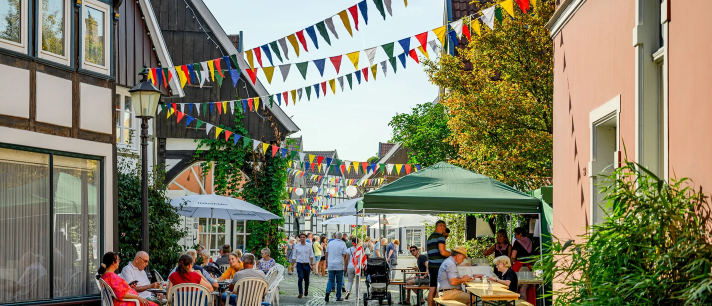 0439.jpg Bunt beflaggte Kopfsteinpflasterstraße mit Menschen in sommerlicher Atmosphäre unter Sonnenschirmen.