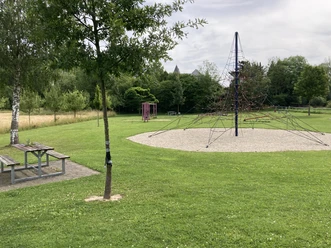 Großer Wasserspielplatz mit Kletterspinne und Picknickbereich, umgeben von grüner Landschaft.