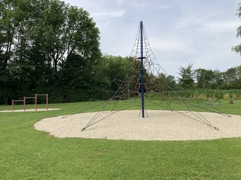 Großes Klettergerüst aus Seilen auf einem runden Kiesplatz, umgeben von grüner Wiese und Bäumen.