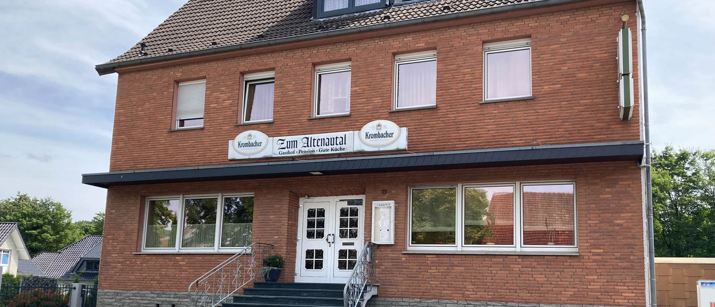 Gasthof im roten Backsteinbau mit überdachter Treppe und weißen Türen, umgeben von Bäumen.