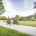 Donauradweg bei Matting Zwei Radfahrer radeln mit E Bikes auf einem asphaltierten Abschnitts des Donauradwegs bei Matting. Im Hintergrund ist die Donau mit den Jurahängen zu sehen.