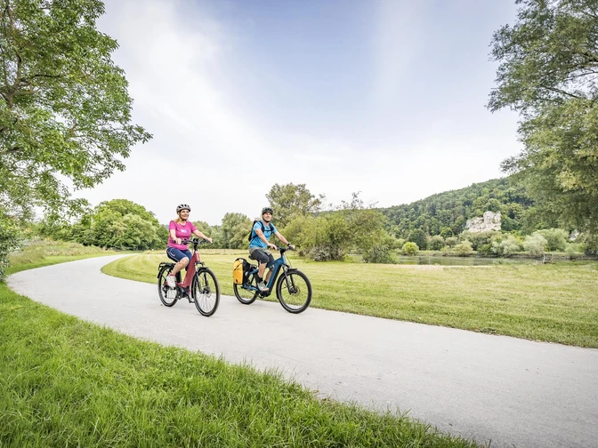 Donauradweg bei Matting Zwei Radfahrer radeln mit E Bikes auf einem asphaltierten Abschnitts des Donauradwegs bei Matting. Im Hintergrund ist die Donau mit den Jurahängen zu sehen.