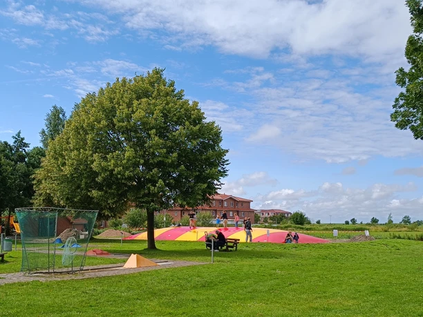 Kinder toben auf einem großen rot-gelben Hüpfkissen auf einer weitläufigen grünen Spielwiese.