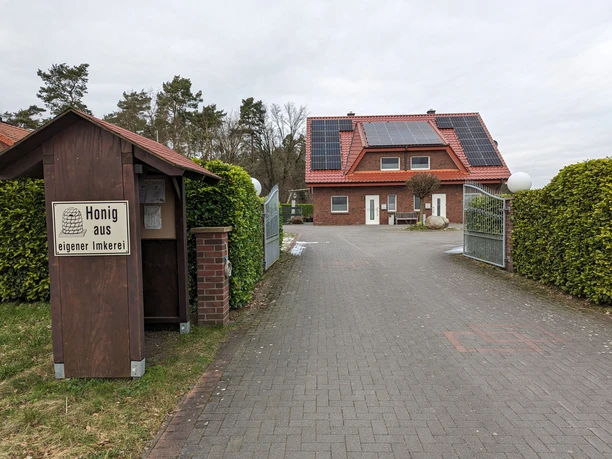 FW_Kurmann_Spelle1©A.Kurmann.jpg Einfamilienhaus mit rotem Ziegeldach und Solaranlage, Einfahrtstor und Holzstand mit Honigverkauf.