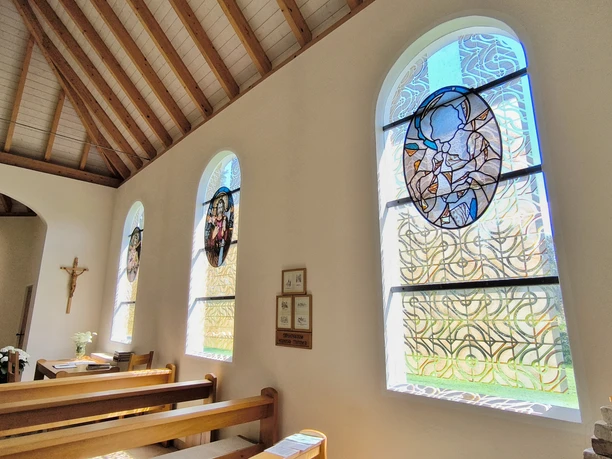 Hövelsenner Kapelle Drei bunte Glasfenster werfen farbenfrohe Lichtreflexe in die Holzkirche mit Holzdeckenbalken.