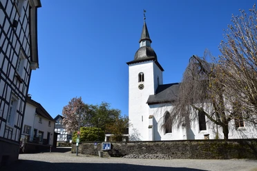 Bonte Kerke in Nümbrecht - Marienberghausen <p>Katholische Kirche mit weißer Fassade und Turm, umgeben von Fachwerkhäusern unter blauem Himmel.</p>