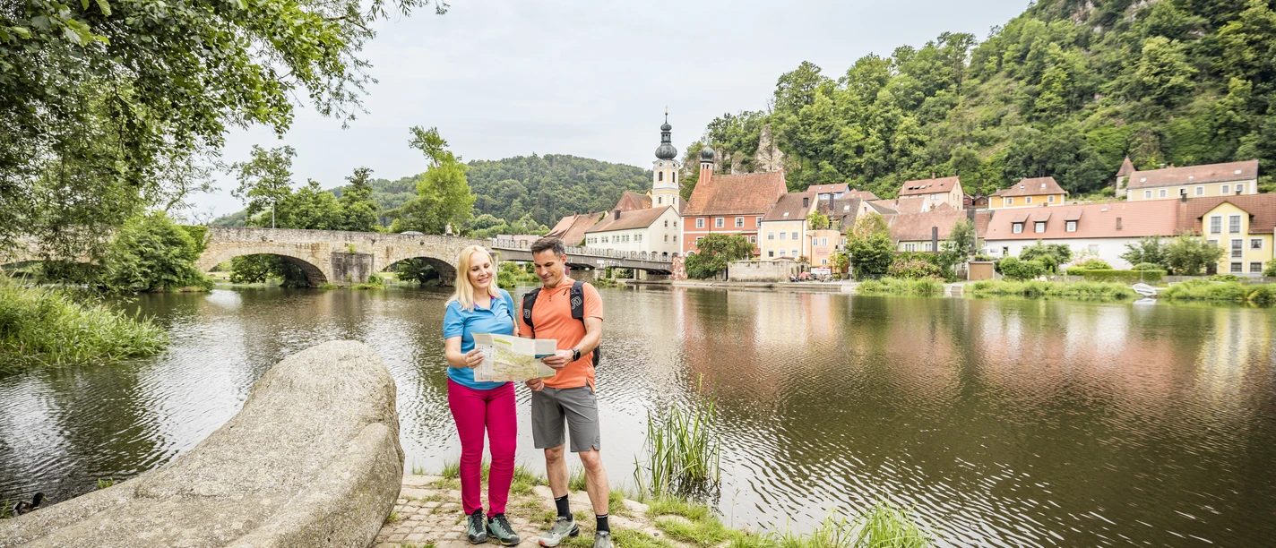 Kallmünz am Naabufer Ein Pärchen steht mit einer Übersichtskarte am Ufer der Naab, im Hintergrund ist der bekannte Künstlerort Kallmünz zu sehen