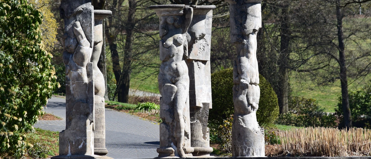 Kurpark in Nümbrecht Kunstvolle Skulpturen aus Stein auf einem pflastersteinernen Weg, umgeben von Bäumen und Sträuchern.