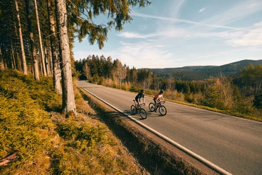 Zwei Rennradfahrer auf einer Waldstraße im Sauerland mit weitem Blick über Hügel.