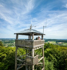 Borgholzhausen-Luisenturm-Teutoburger-Wald-Tourismus-D-Ketz-056.jpg Holzturm in grüner Landschaft mit Ausblick, zwei Personen geniessen die Aussicht unter blauem Himmel.