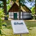 Fachwerk-Gebäude im Grünen hinter einer Infotafel, umgeben von Bäumen und Schatten.