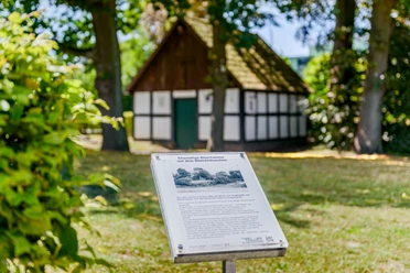 _BBB7326.jpeg Fachwerk-Gebäude im Grünen hinter einer Infotafel, umgeben von Bäumen und Schatten.