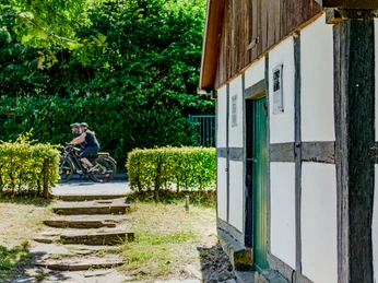 _BBB7372.jpeg Radfahrer fahren auf einem sonnigen Weg an einem alten Fachwerkhaus mit Holztüren und grüner Umgebung vorbei.