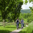 Zwei Personen fahren mit Fahrrädern auf einem grünen Waldweg, umgeben von Bäumen und Wiese.