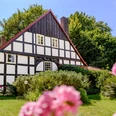 Fachwerkhaus mit weißer Fassade und dunklen Balken, umgeben von üppigem Grün und rosa Blumen.