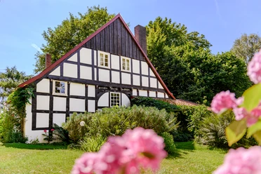 _BBB7459.jpeg Fachwerkhaus mit weißer Fassade und dunklen Balken, umgeben von üppigem Grün und rosa Blumen.