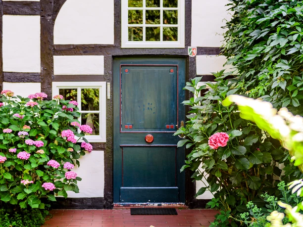 _BBB7469.jpeg Historisches Fachwerkhaus mit blaugrüner Tür, umgeben von blühenden Hortensien und dichtem Grün.