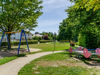 Grüner Spielplatz mit Schaukel, Wipptier und Bänken, umgeben von Bäumen und Wohnhäusern im Hintergrund.