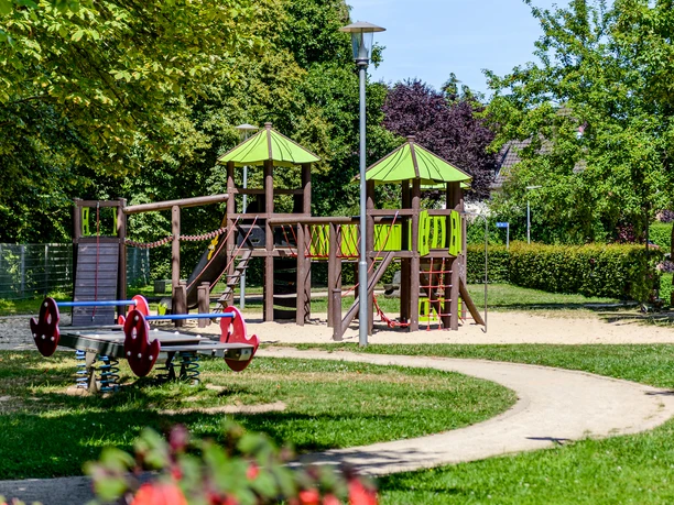 Ein Spielplatz mit Klettergerüst, Rutsche, Wippe und umgebenden Bäumen bei sonnigem Wetter.